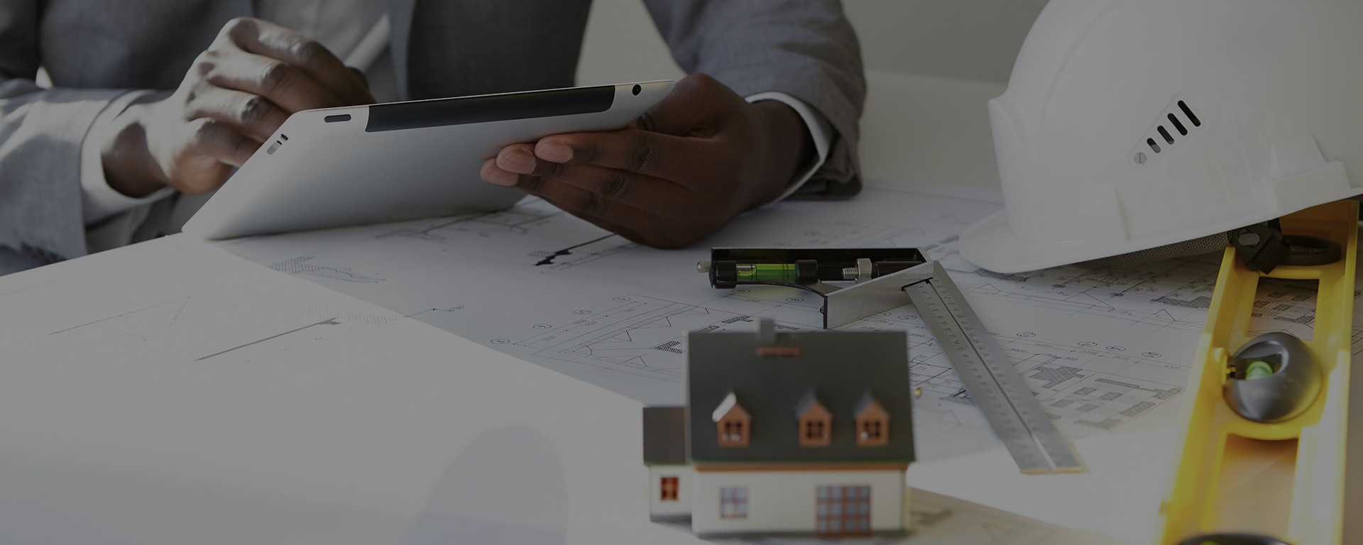Cropped shot of dark-skinned contractor holding touch pad, entering data while working on new housing project, sitting at desk with drawings, scale model house, blueprint rolls, ruler and helmets